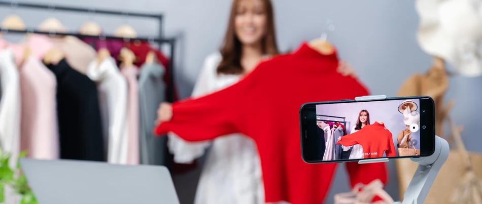A smartphone sits on a tripod and records a person holding up and displaying a red shirt.