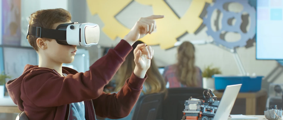 A young boy in a classroom environment with a VR headset on with his fingers pointing out in front of him while other student study on computers behind him