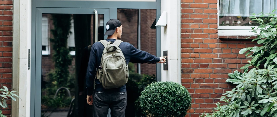 A man wearing a backwards baseball hat with a backpack facing the glass front of an office building ringing the doorbell with his right index finger